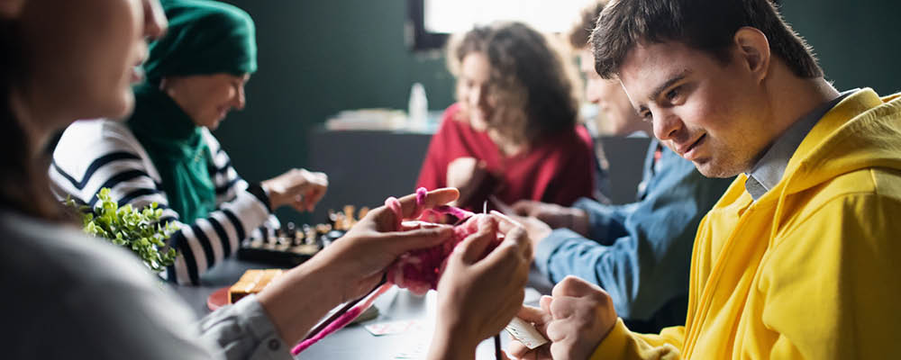 Direct Support Professionals and their clients gathered around a table, knitting and playing chess.